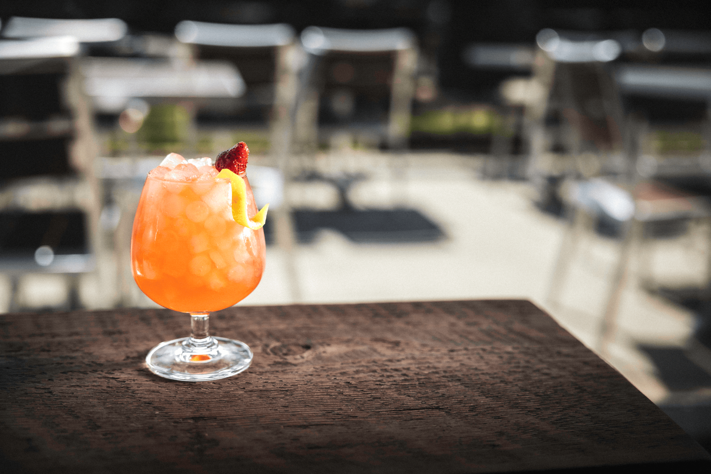 A vibrant orange cocktail with ice, garnished with a lemon twist and a raspberry, stands on a wooden table.