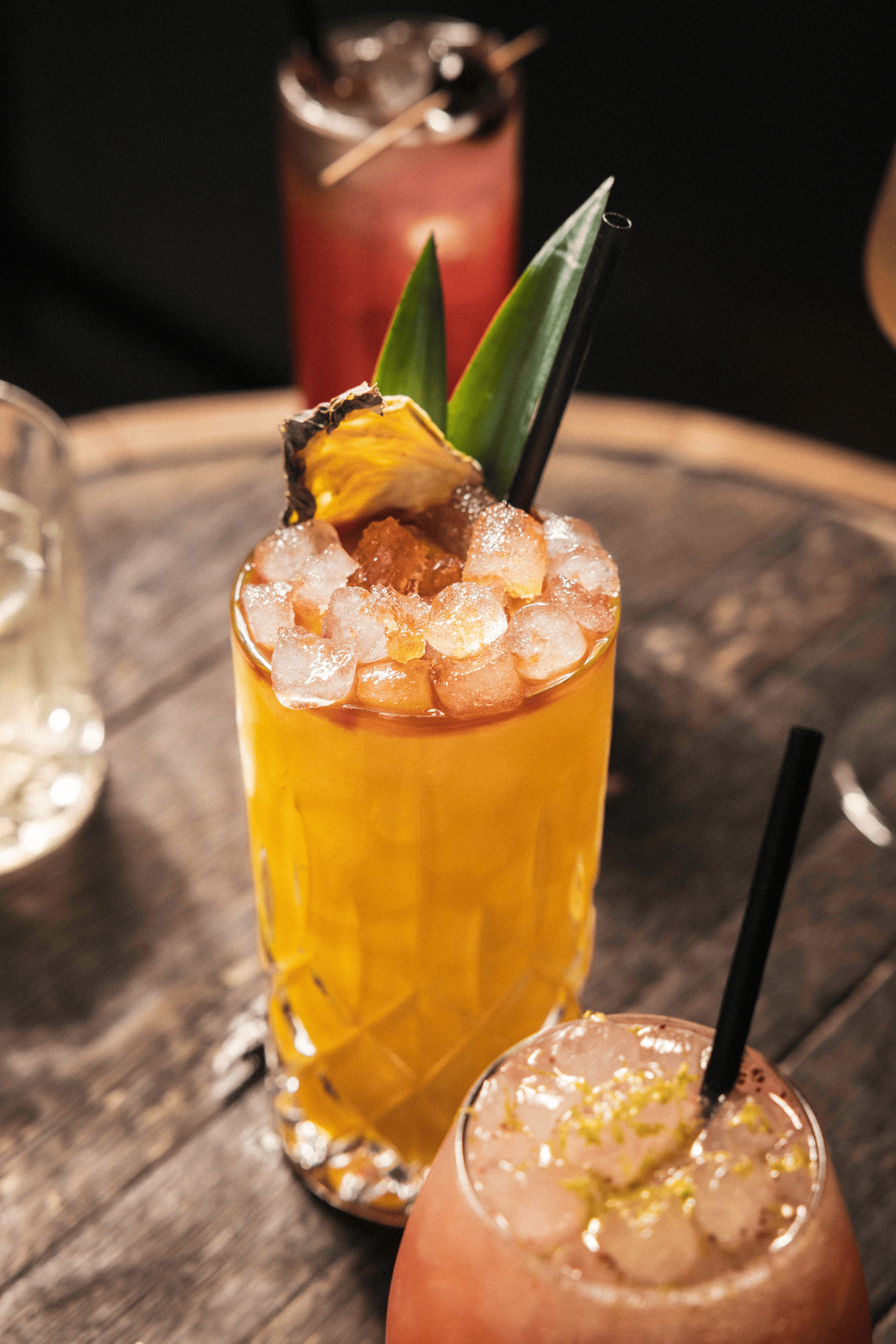 A vibrant yellow cocktail garnished with a pineapple slice and green leaves sits atop a rustic wooden table, glistening with ice.