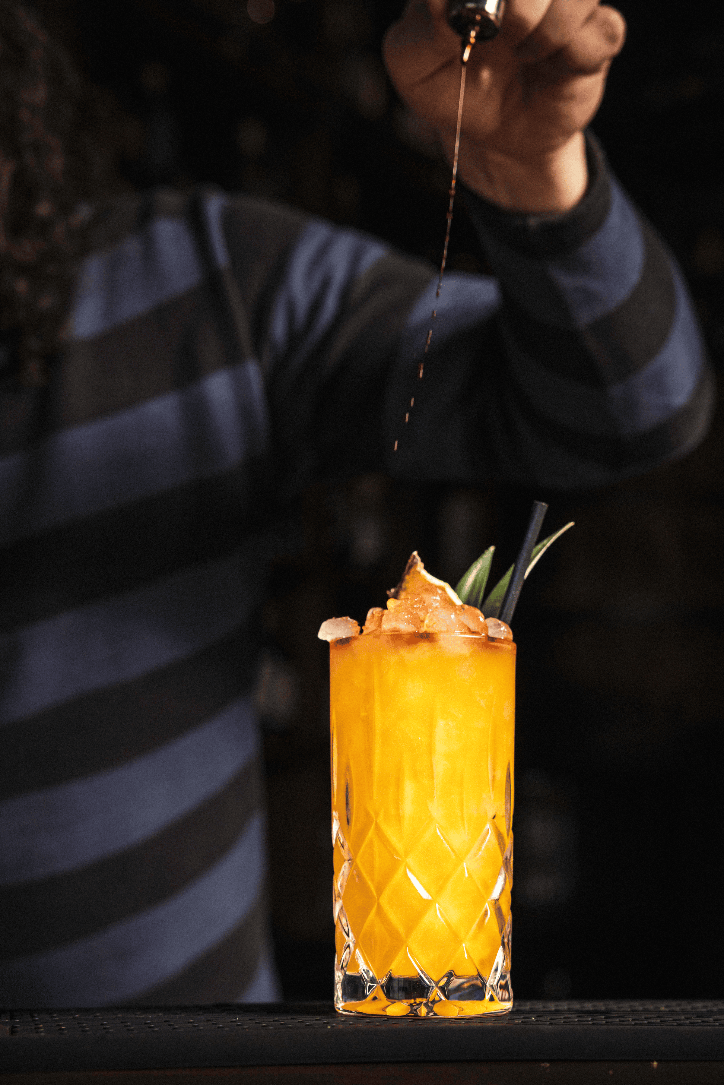A vibrant orange cocktail filled with ice is garnished with a slice of fruit and green foliage, being expertly crafted.