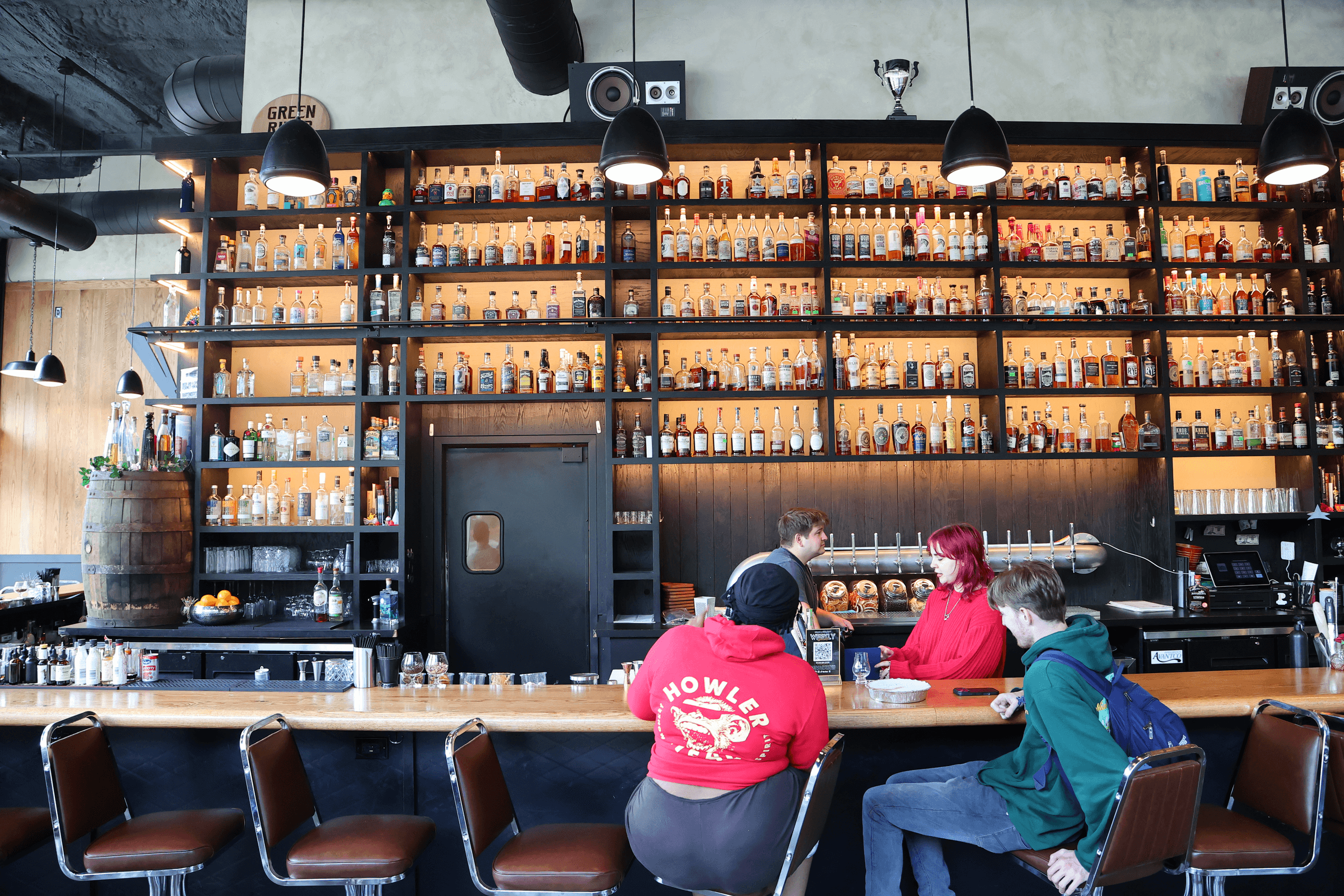A stylish bar features a towering shelf lined with colorful bottles, while patrons lounge on retro stools, enjoying cocktails.