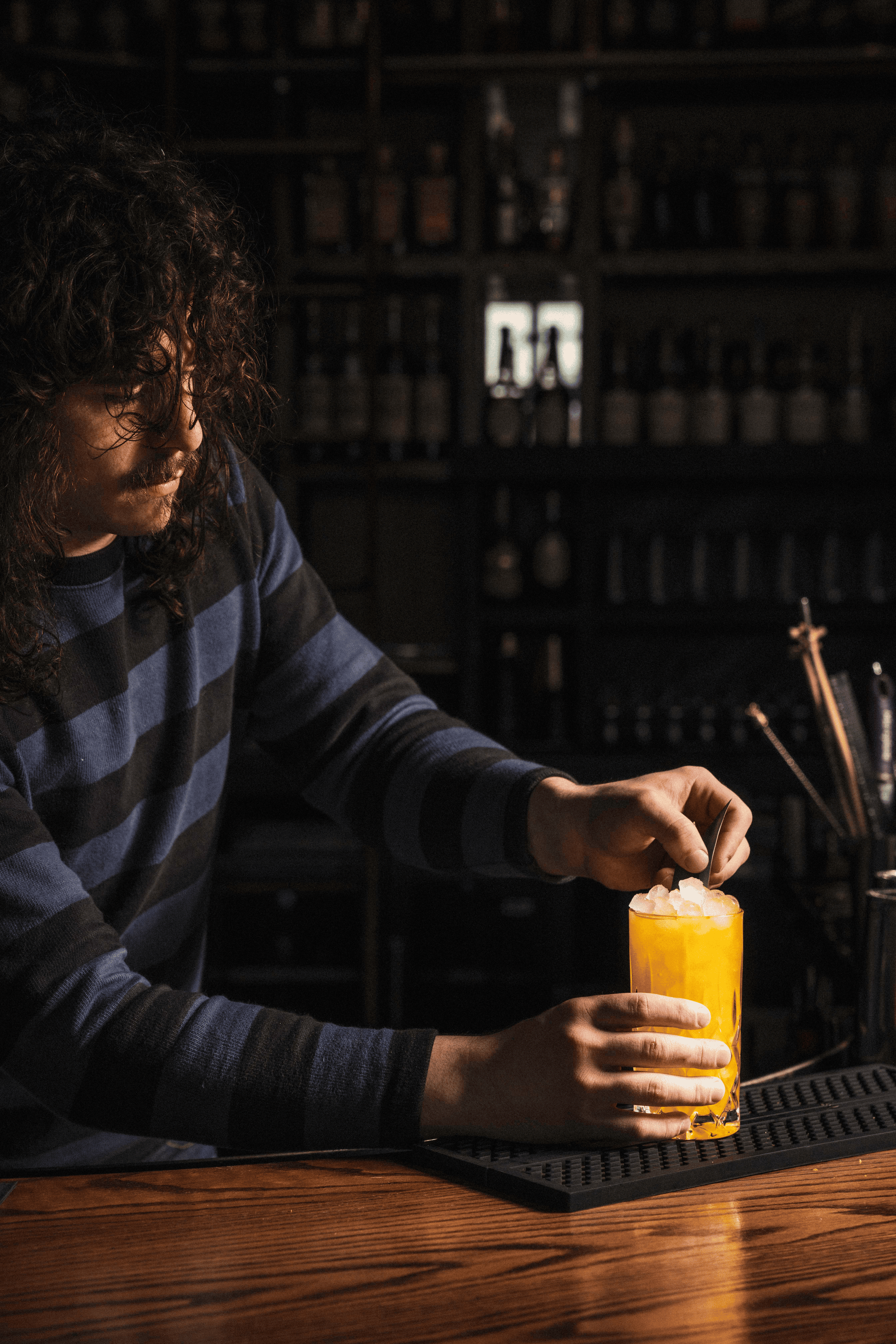 A bartender carefully garnishes a bright orange cocktail filled with ice, surrounded by a backdrop of liquor bottles.
