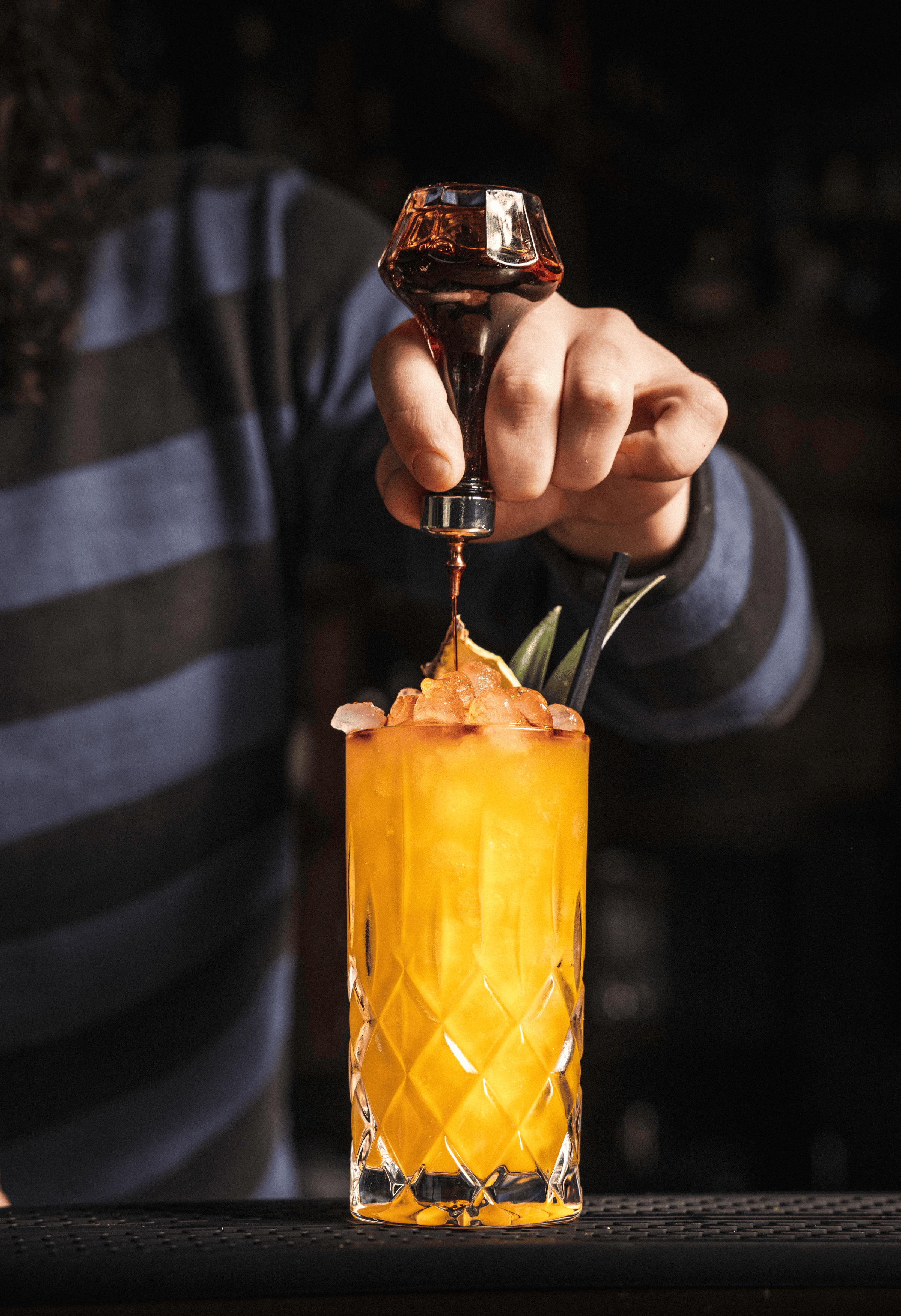 A bartender drizzles dark syrup over a vibrant orange cocktail topped with crushed ice and garnished with green herbs.